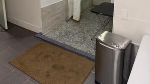Une salle de bain avec une douche, des murs carrelés, un rideau blanc, une poubelle en métal et un paillasson marron.
