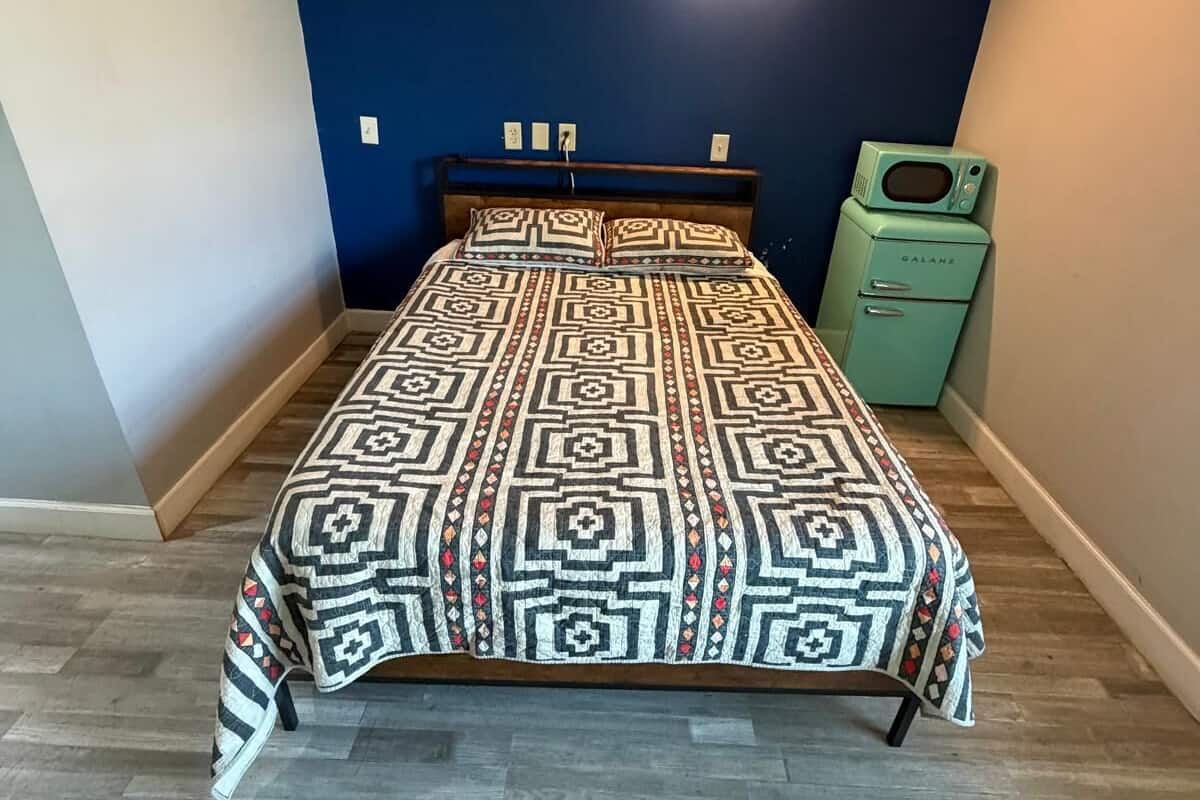 A bedroom with a patterned bedspread, a wooden headboard, a small green appliance, and a wall-mounted light fixture.
