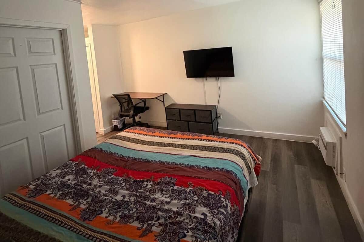A bedroom with a large bed, a desk, a chair, a TV, and a window with blinds.