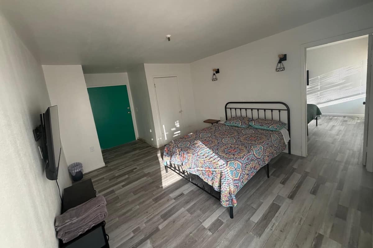 A bedroom with a bed, a green door, a small table, and a TV mounted on the wall.