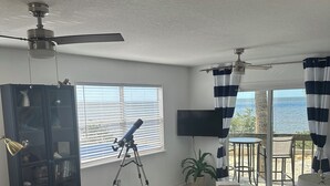 Sala de estar com um telescópio, ventilador de teto, estante de livros e vista para o oceano através de grandes janelas.