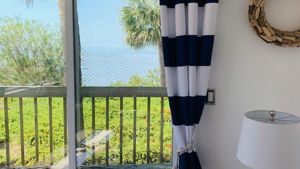 Quarto com uma grande janela, vista para um deck de madeira e palmeiras, cortina, luminária e uma grinalda decorativa.