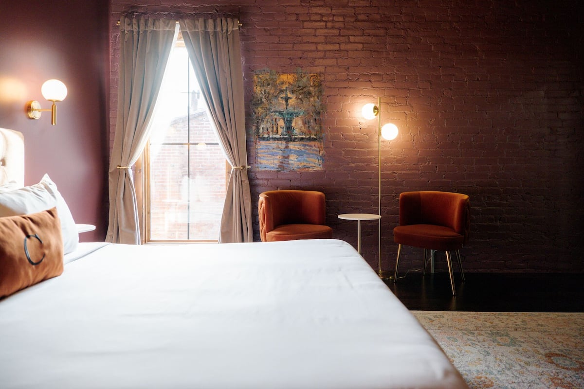 A hotel room with a large bed, two armchairs, a small round table, and a painting on the brick wall.