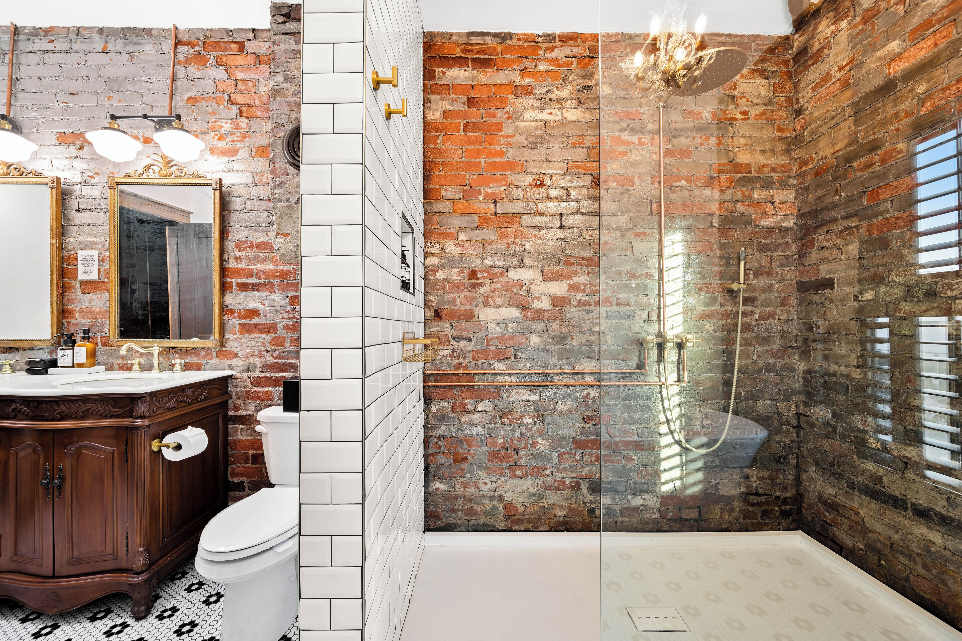 Un baño con inodoro blanco, tocador de madera y zona de ducha con mampara y pared de ladrillo.