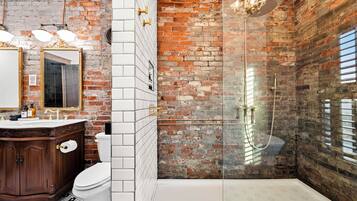Un baño con inodoro blanco, tocador de madera y zona de ducha con mampara y pared de ladrillo.
