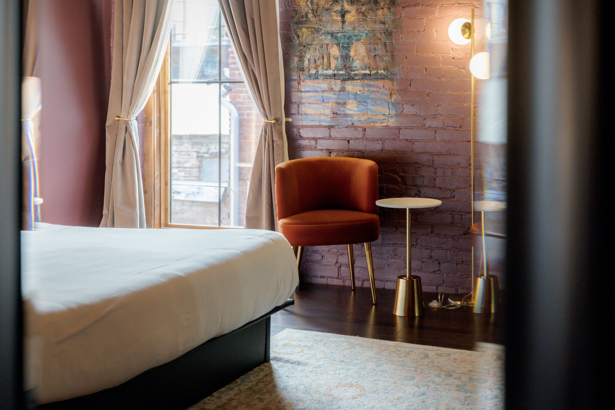 A hotel room with a bed, an orange armchair, a small round table, and a wall with a brick pattern.