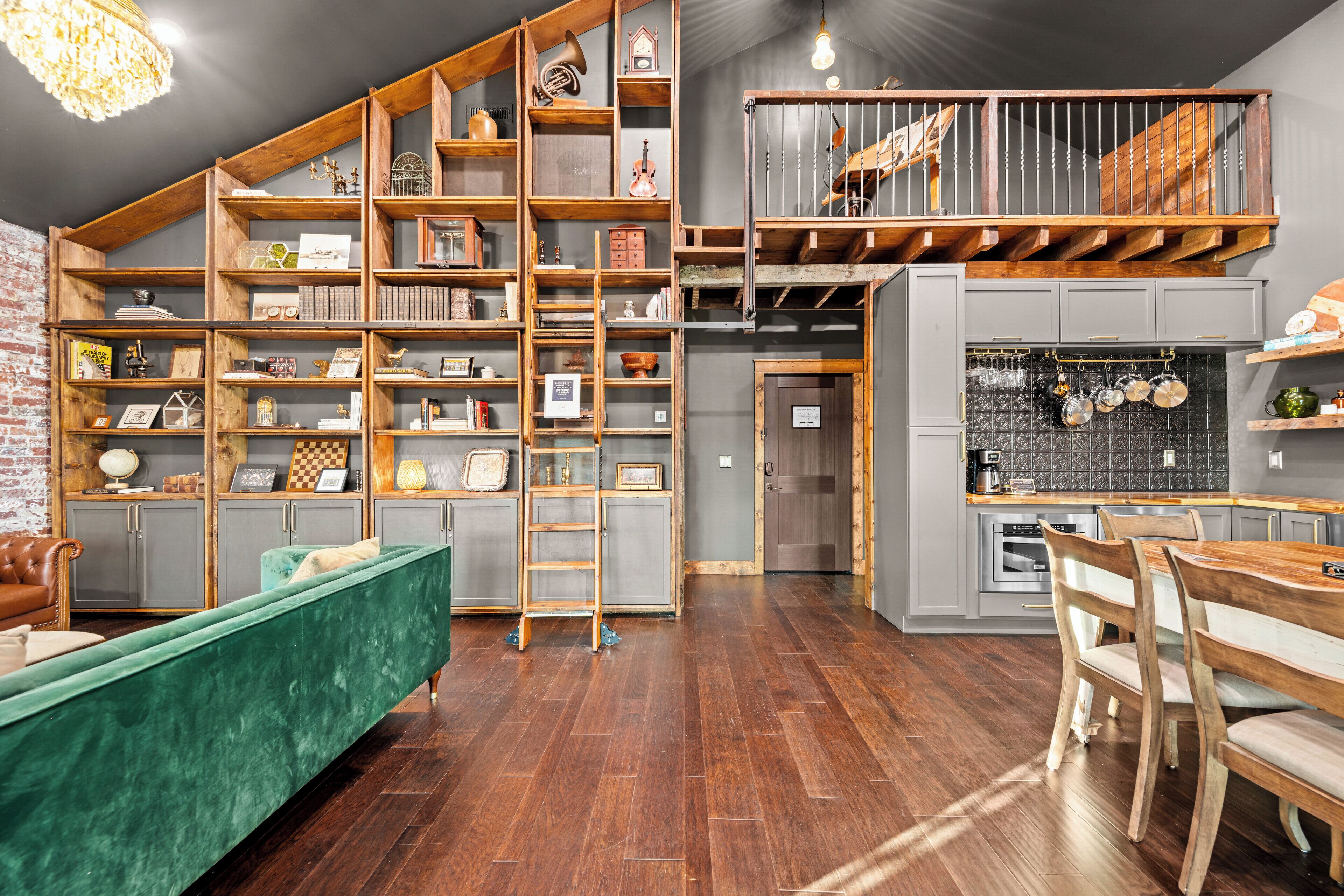Amplio espacio tipo loft con cama superior, estanterías de madera llenas de libros y objetos decorativos, un sofá verde y una zona de comedor con sillas y mesa de madera.