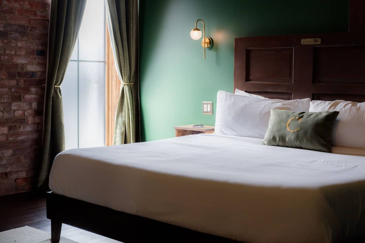 A neatly made bed with white linens and a green pillow, a wooden headboard, a nightstand with a lamp, and a brick wall with a window and curtains.