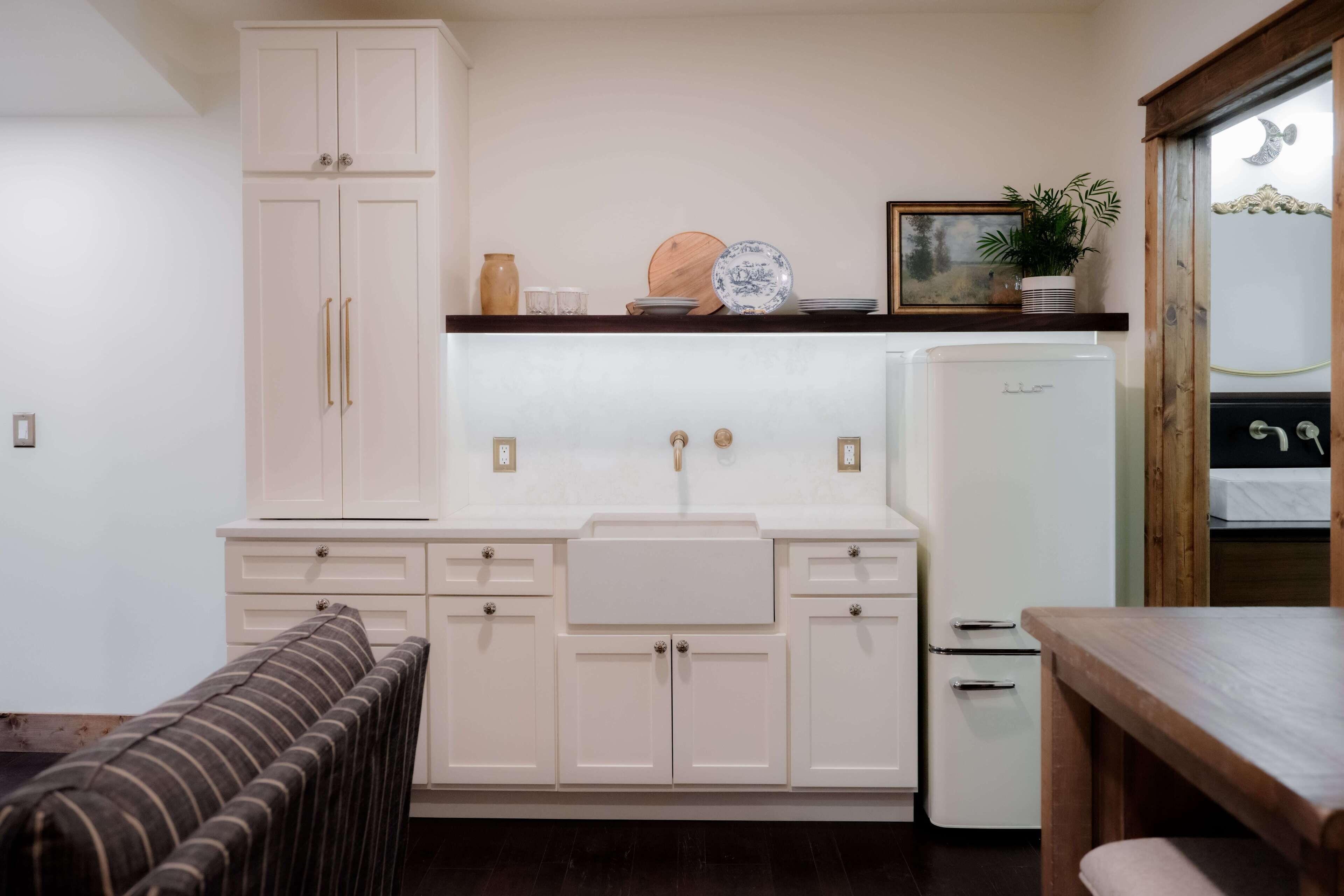 A compact kitchen with white cabinets, a built-in sink, and a small refrigerator. There is a wooden table and a bench in the foreground.