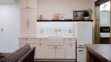 A compact kitchen with white cabinets, a built-in sink, and a small refrigerator. There is a wooden table and a bench in the foreground.