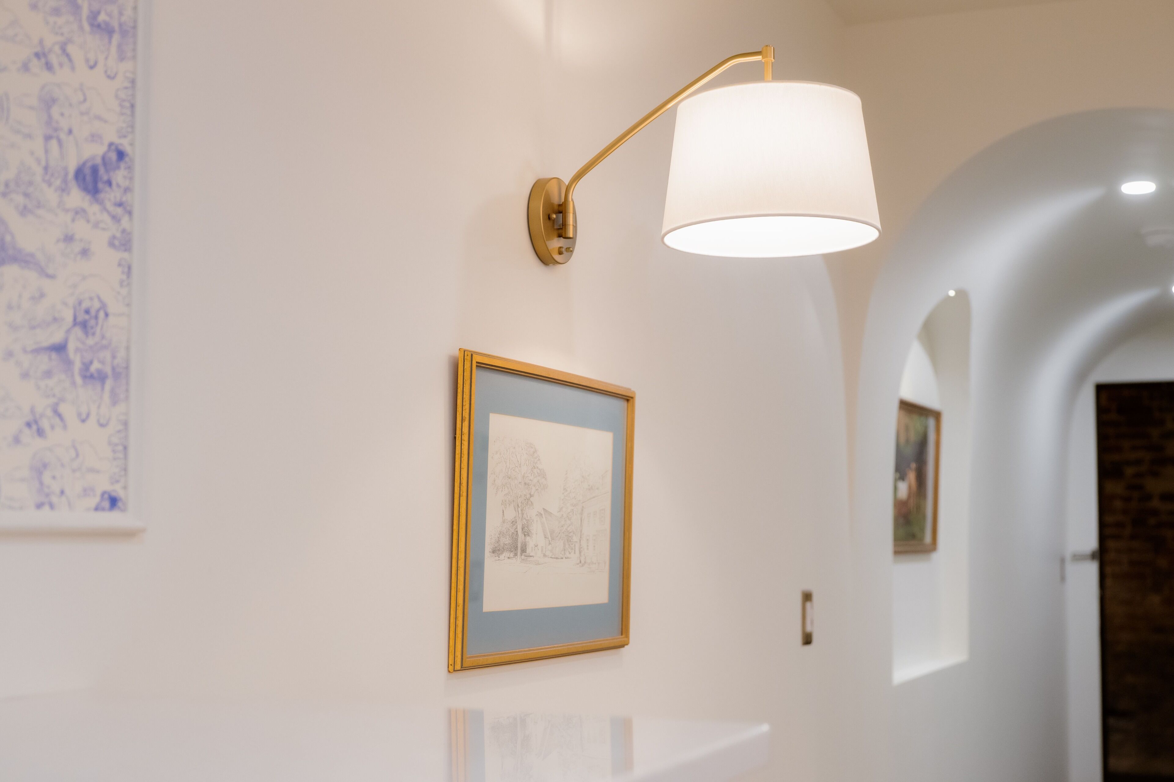 A hallway with a framed artwork on the wall, a wall-mounted lamp, and a reflective surface.