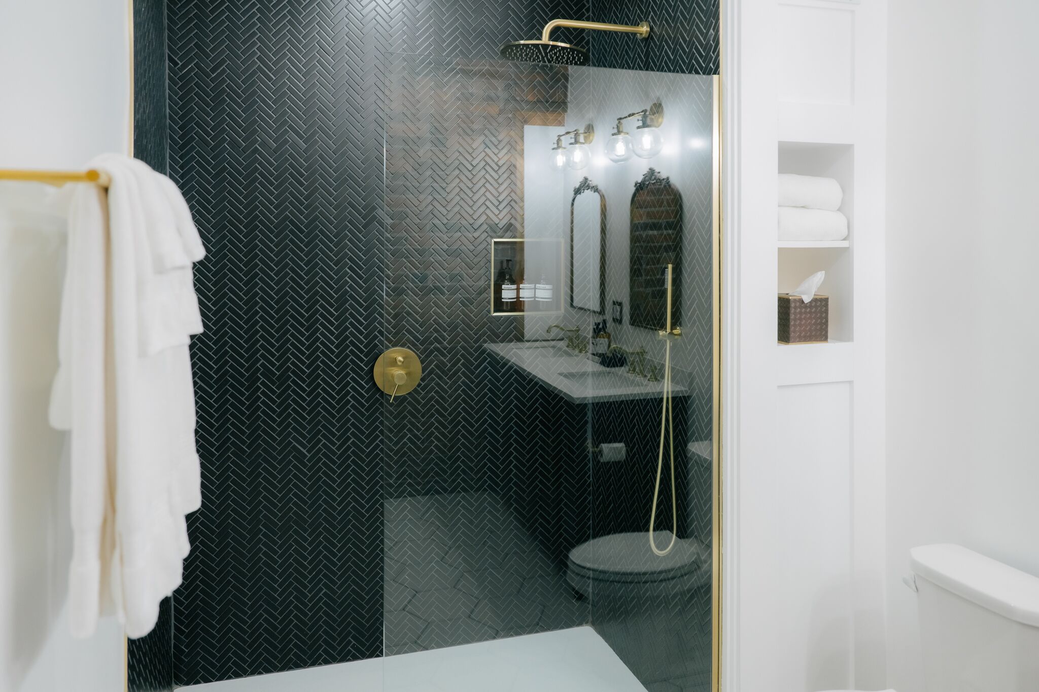 A modern bathroom with a black herringbone tile wall, a glass shower enclosure, a white sink, and a toilet.