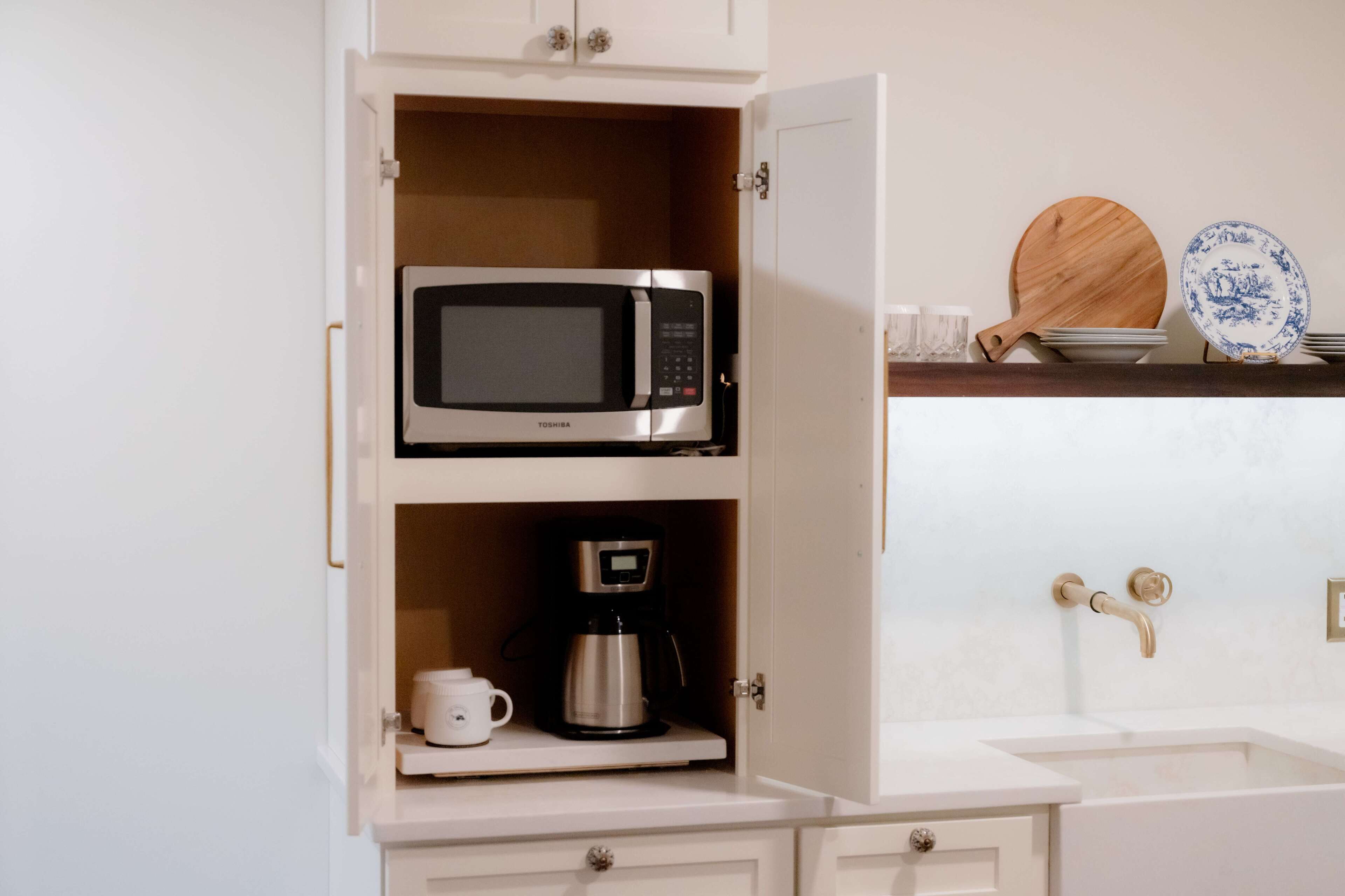 A kitchen with a microwave and coffee maker on a shelf, a sink with a gold faucet, and wooden cutting boards and plates on the counter.