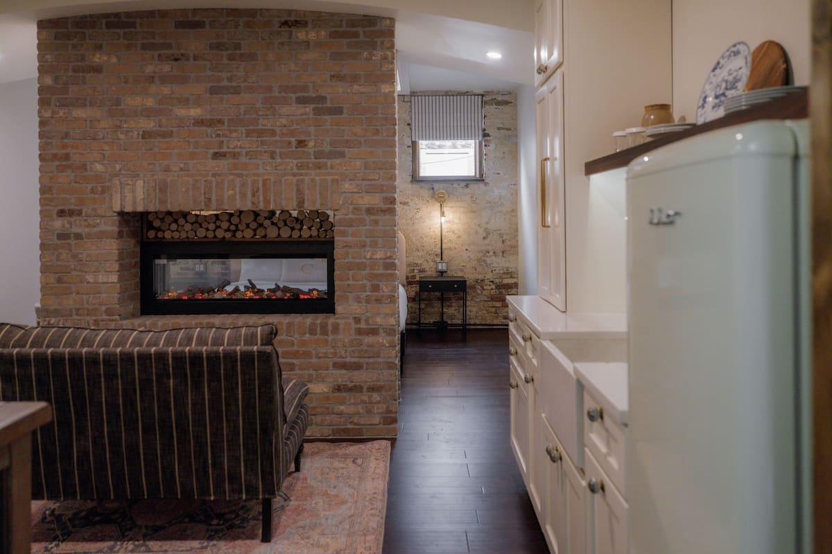 A cozy living room with a brick fireplace, a sofa, and a dining area with a refrigerator.