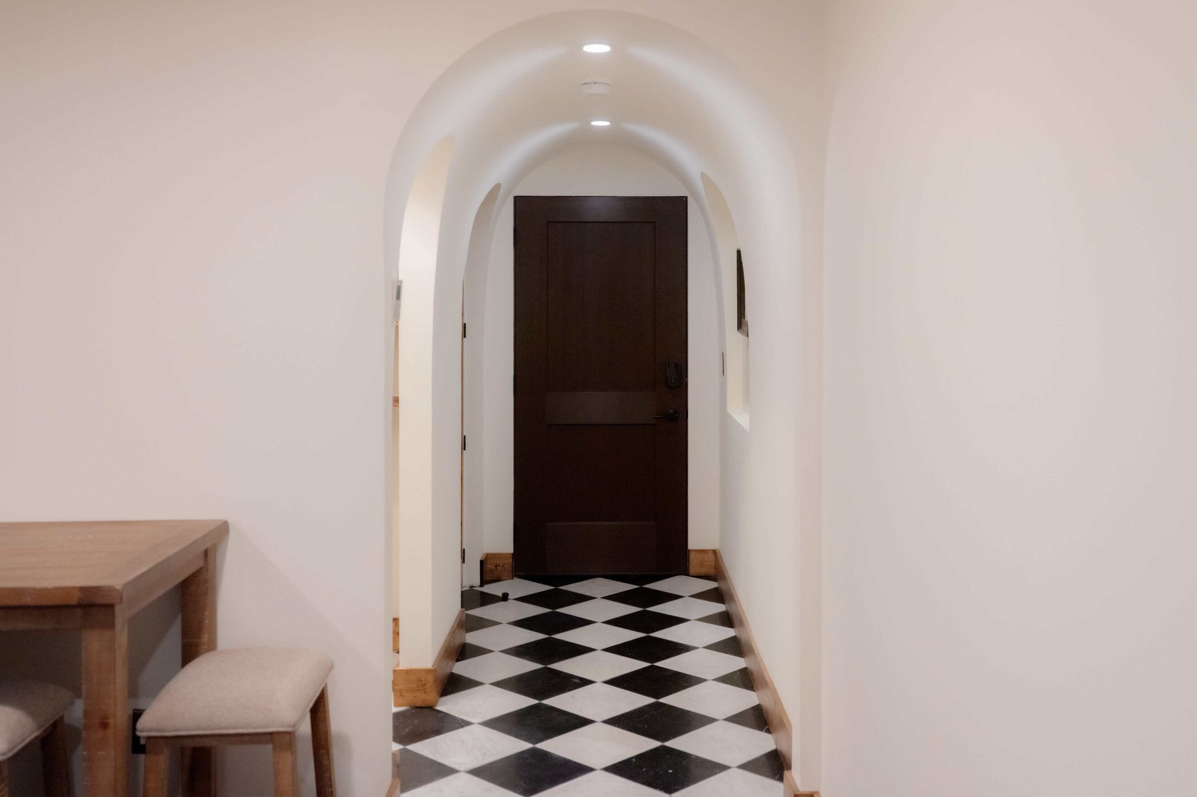 A narrow hallway with a checkered floor, a wooden table with chairs, and a door at the end.