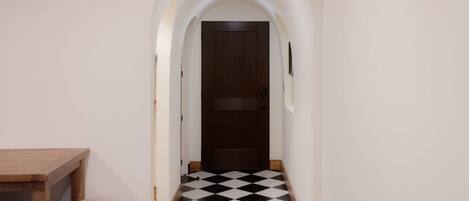 A narrow hallway with a checkered floor, a wooden table with chairs, and a door at the end.