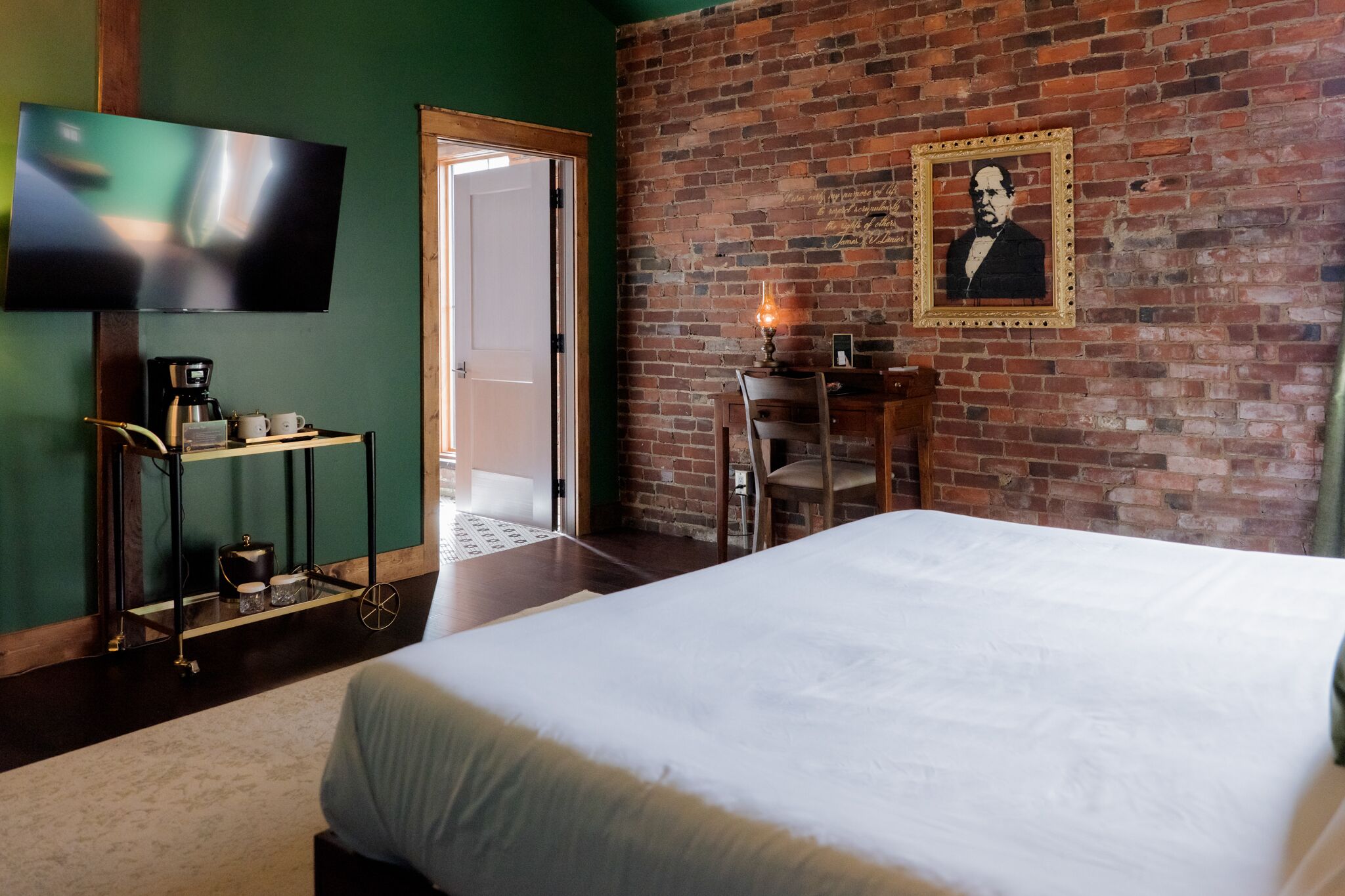 Habitación de hotel con pared de ladrillo, televisor de pantalla plana, cama, escritorio con silla y pequeña mesa con lámpara.