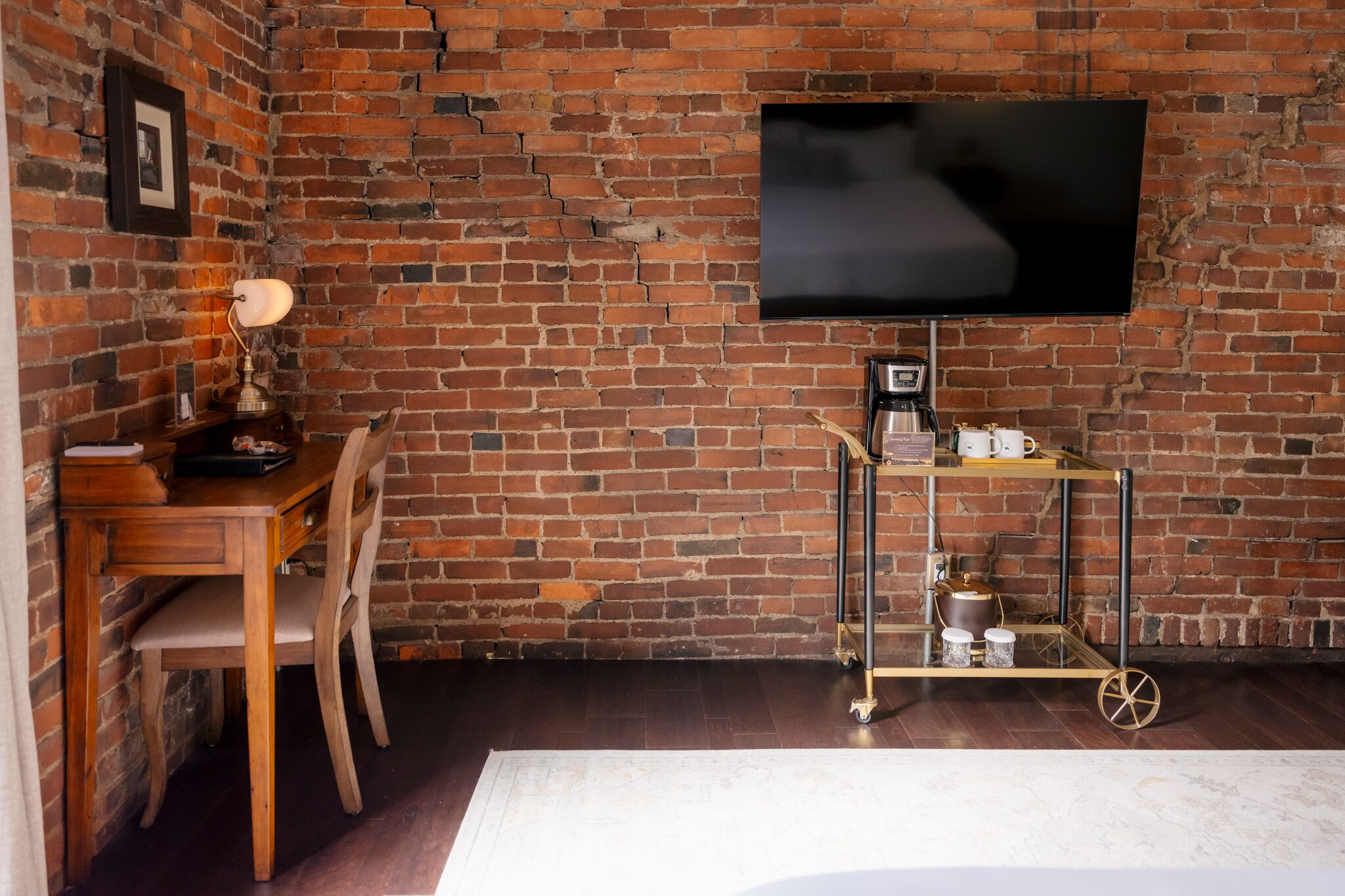 A room with a brick wall, a wooden desk, a chair, a TV mounted on the wall, and a cart with items.