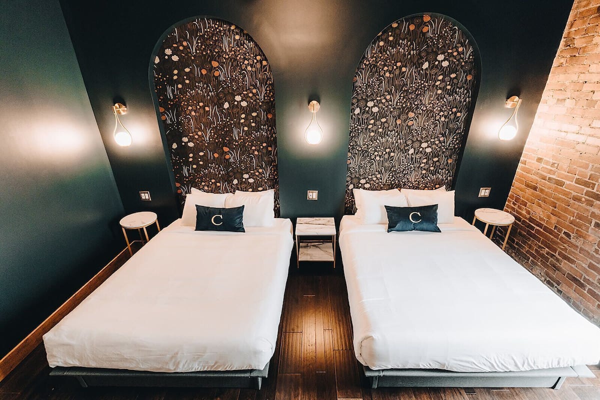 A double room with two beds, each with a pillow marked with a 'C', a small table, and a wall-mounted light fixture.