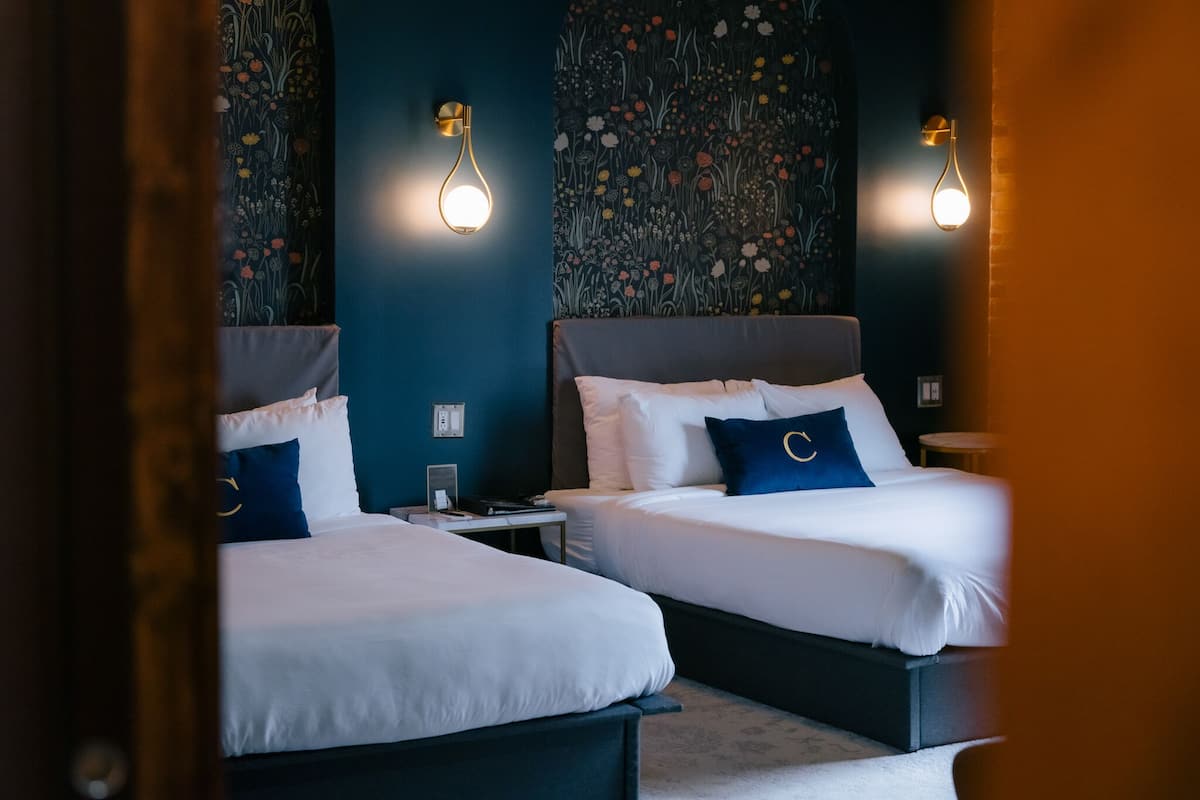 A hotel room with two beds, each with white bedding and blue pillows, and a floral-patterned wall.