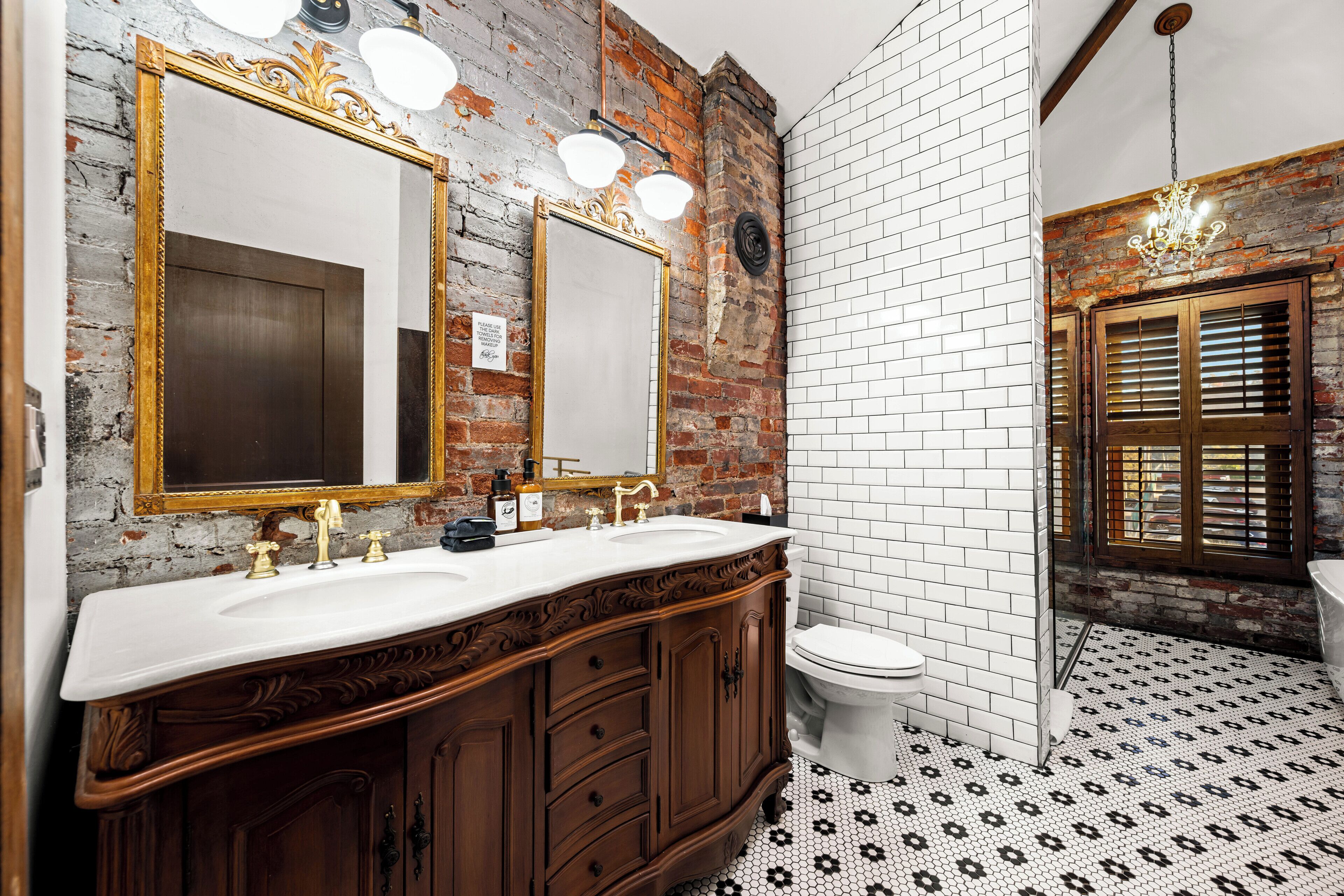 Un baño con pared de ladrillo rústica, piso de baldosas blancas y tocador de madera con dos lavamanos.