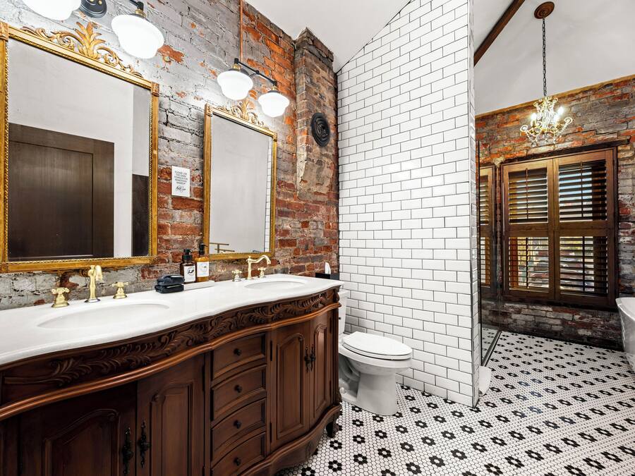 Un baño con pared de ladrillo rústica, piso de baldosas blancas y tocador de madera con dos lavamanos.