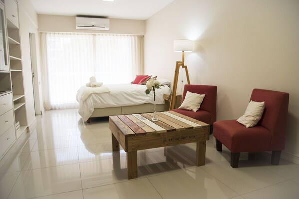 Une chambre d’hôtel avec un lit, une table basse en bois, deux fauteuils rouges, une lampe sur pied et une commode blanche.