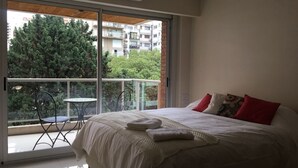 Een slaapkamer met een groot bed, een balkon met een tafel en stoelen, en uitzicht op gebouwen en bomen.