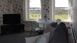A living room with a TV, a sofa, a table with a vase, and two windows with a view of a park and buildings. - Special seafront apartment with stunning view in characterful Victorian building (Portsmouth)