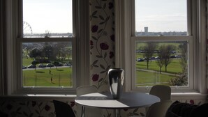 A room with two large windows, a white table, and a chair. Outside, there is a park with cars and a Ferris wheel. - Special seafront apartment with stunning view in characterful Victorian building (Portsmouth)