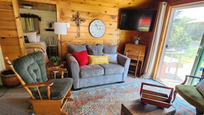 Ein gemütliches Blockhäuschen-Interieur mit Holzbalkendecke, einer Couch, einem Sessel, einem kleinen Tisch und einem großen Fenster.