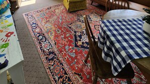 Ein gemütliches Zimmer mit Deckenventilator, einem großen Teppich, einem Tisch mit kariertem Tuch, einem braunen Sofa und einem gelben Sessel.