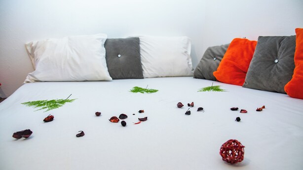 A bed with white bedding, grey and orange pillows, and scattered dried leaves and a red spherical object.