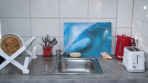 A kitchen with a sink, a toaster, a red kettle, and a painting of a wave.