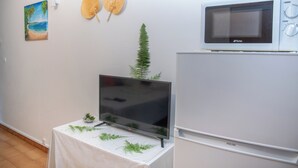 A room with a flat-screen TV on a table, a white refrigerator, a microwave, a painting of a beach, and two woven hats on the wall.