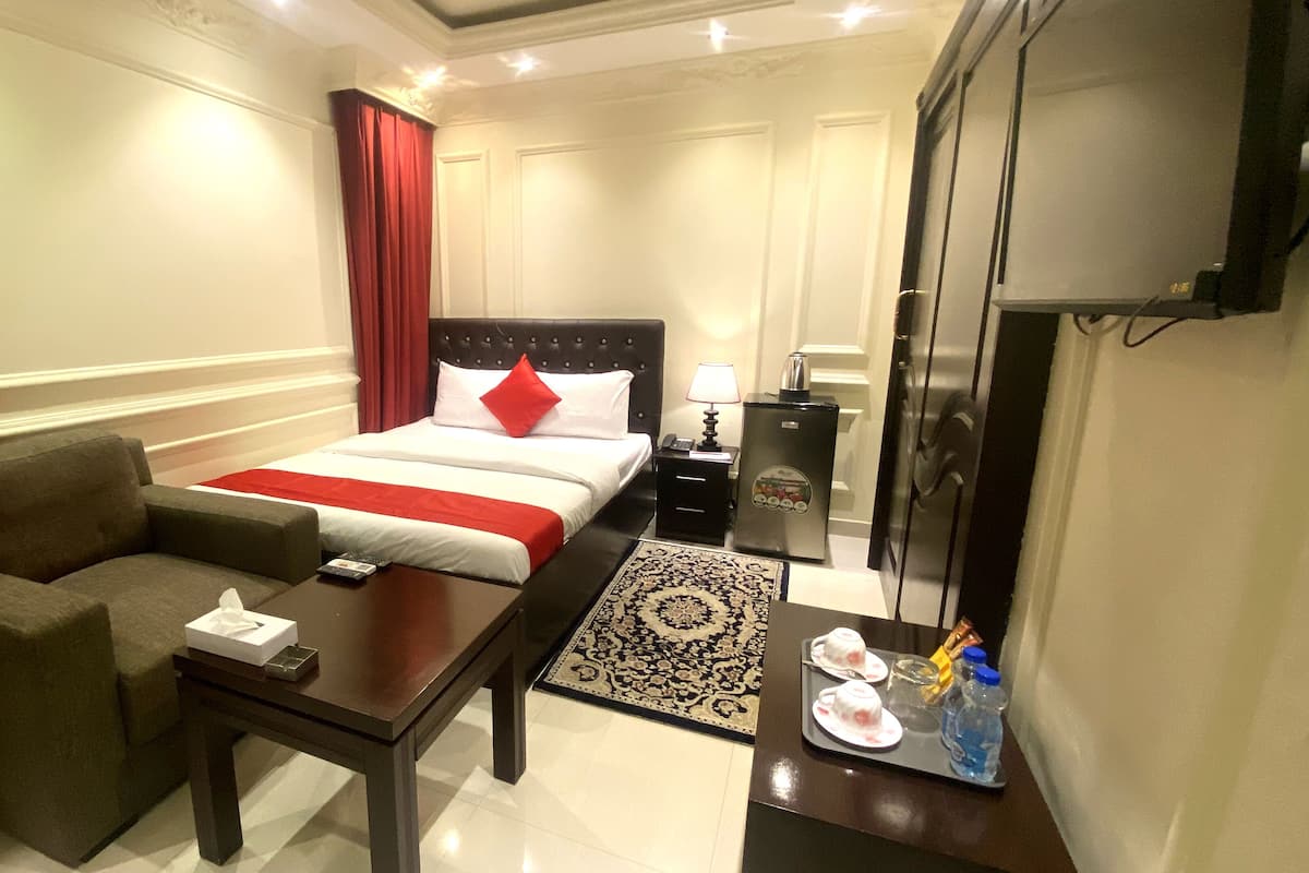 A hotel room with a bed, a sofa, a table, a TV, a refrigerator, and a tray with cups and a bottle.