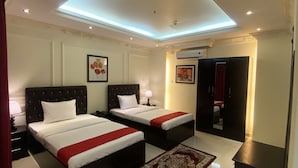 Une chambre d’hôtel avec deux lits, une couvre-lit rouge et blanche, une table de chevet avec une lampe, un tableau encadré au mur, un climatiseur et un miroir dans un coin.