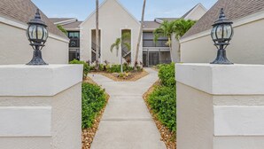 A pathway leading to a building with a modern design, featuring a central entrance flanked by two tall palm trees and decorative street lamps. - Luxury Condo in Gated Golf Community close to Beaches (Fort Myers)