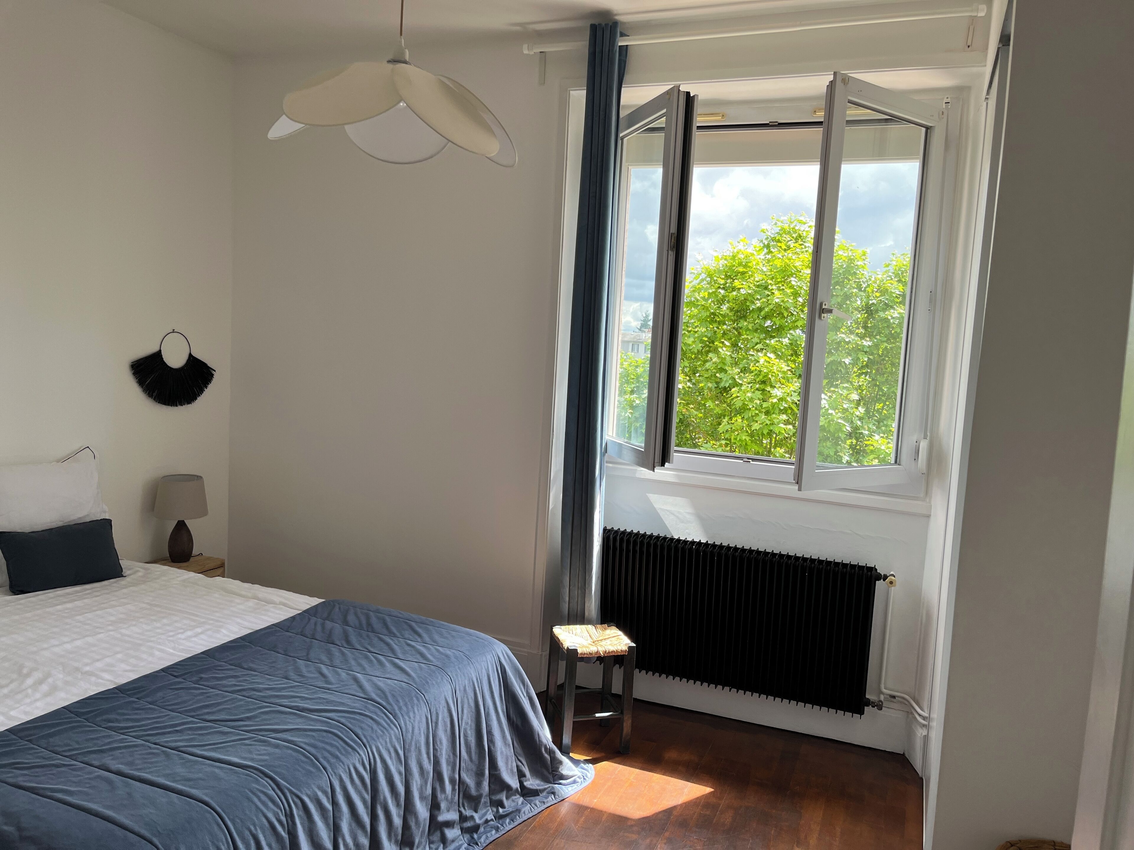 A bedroom with a bed, a nightstand, a lamp, a radiator, and a window with a view of greenery.