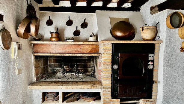 A rustic kitchen with a brick oven, hanging pots, and shelves with dishes.