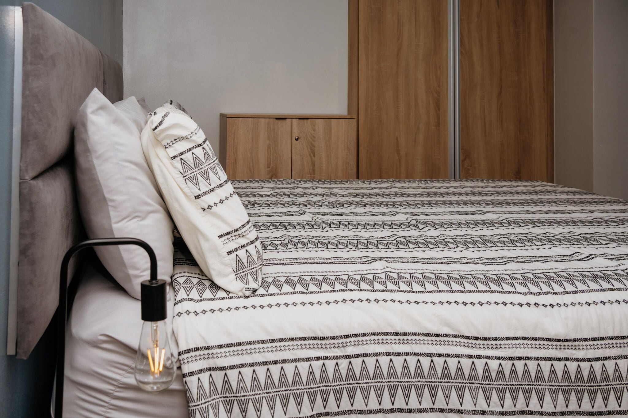A bed with patterned bedding, a bedside lamp, and a wooden wardrobe in the background.
