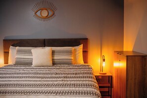 A neatly made bed with patterned bedding, a headboard, and a wall-mounted eye-shaped light fixture. - Amazing Blue Venao, Unique & Cozy 2BR Condo — 3 min from the beach! (Provincia de Los Santos)