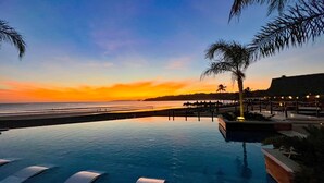 Vista a la piscina con un atardecer sobre el océano, palmeras y una playa.