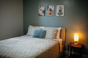 Una cama bien hecha con cubrecolcha acolchado, almohada azul y una lámpara de noche sobre una mesita de madera.