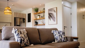 Een moderne woonkamer met een bruine bank, gestreepte kussens, een hanglamp, een keuken op de achtergrond en een deur aan de rechterkant.