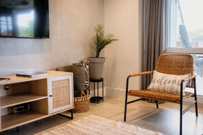 Una sala de estar con un televisor montado en la pared, un sillón de mimbre con cojín, una mesita con una planta y un mueble con estantes.