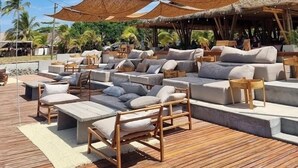 Un área de descanso frente a la playa con asientos de madera, sombrillas de paja y vista al océano.