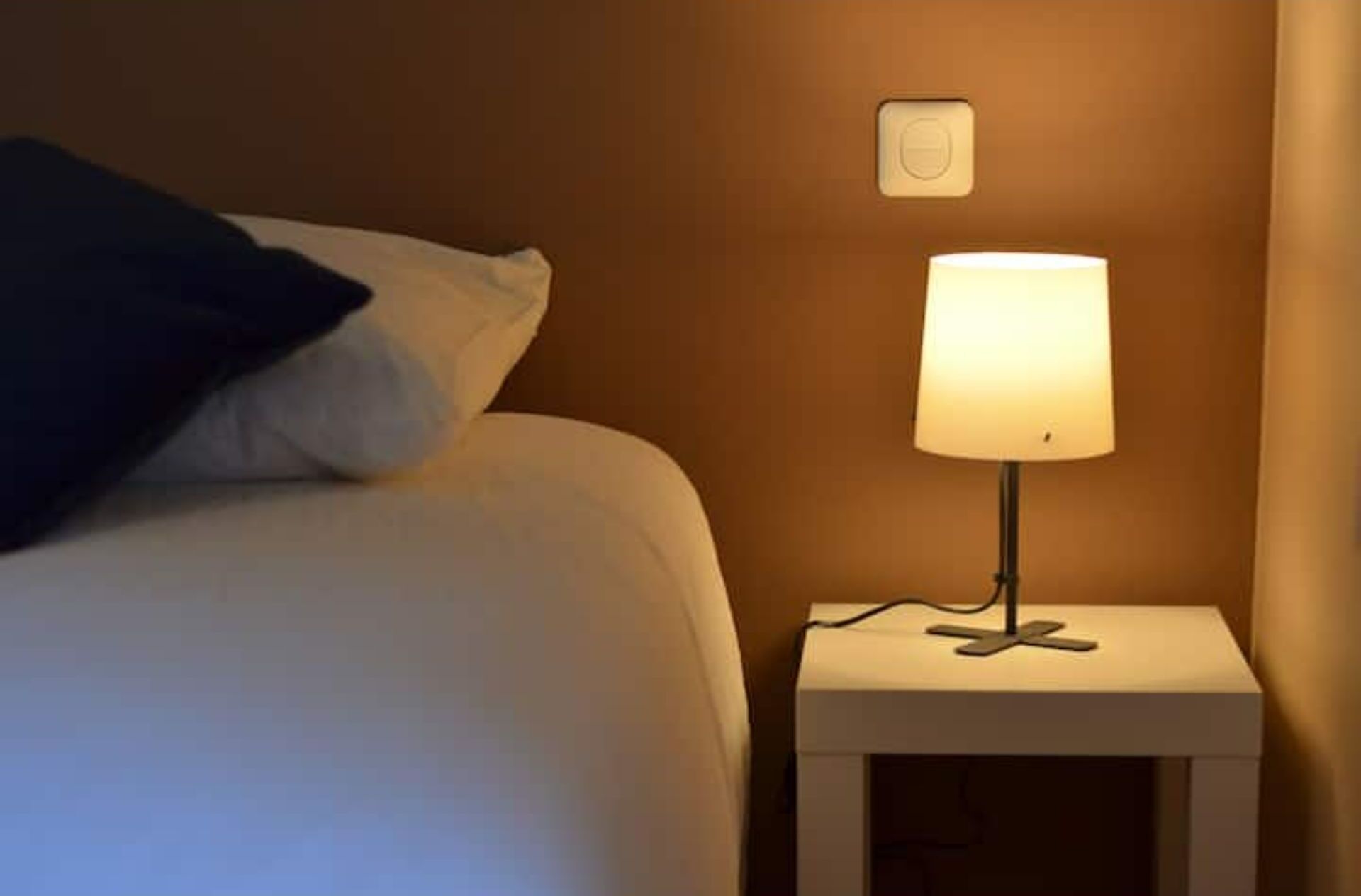 A bed with a white pillow and a dark blue throw, a bedside table with a lamp, and a wall switch.