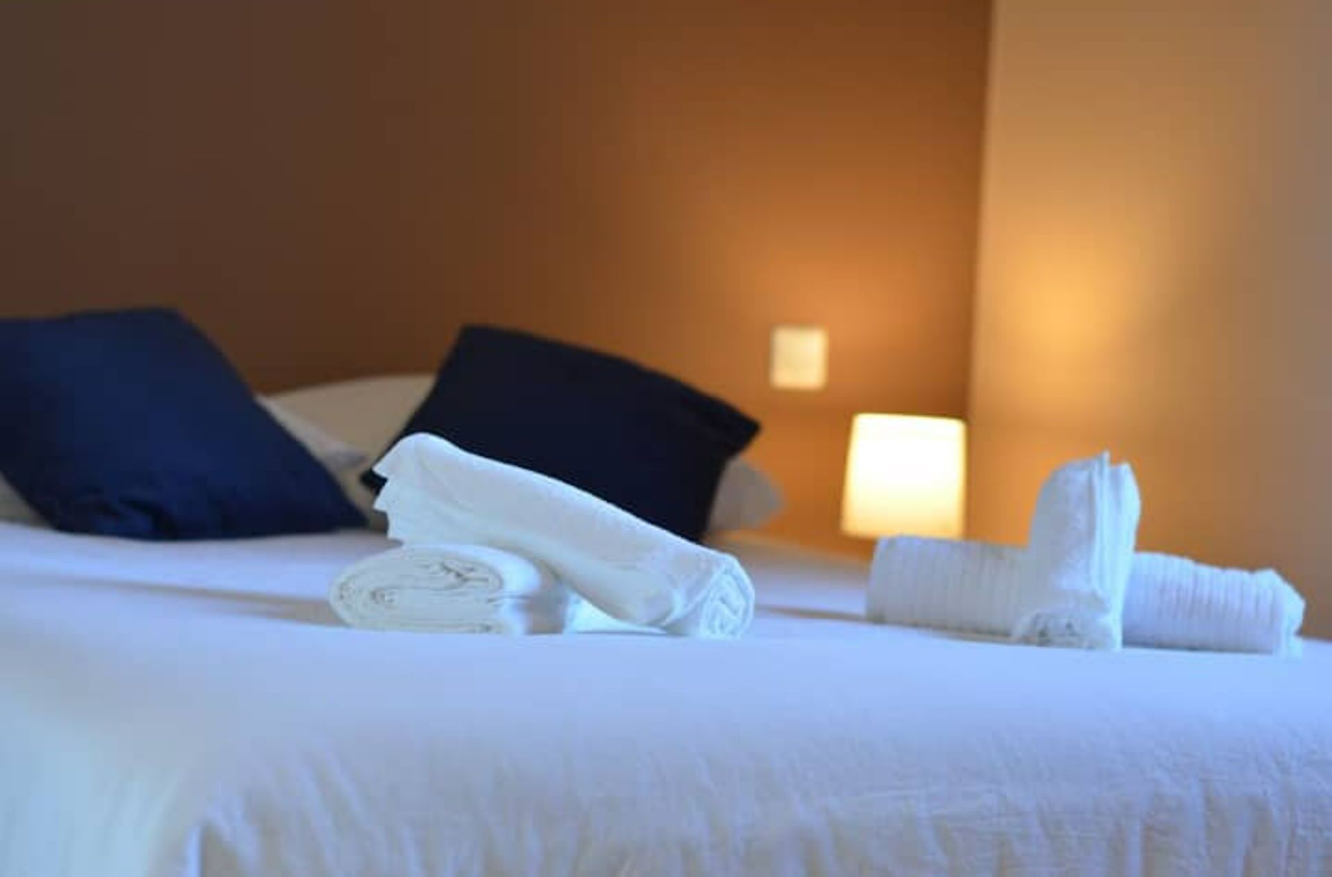 A neatly made bed with white linens, two dark blue pillows, and a folded white towel on the bed.