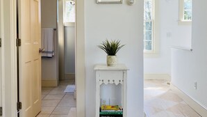 A hallway with a wooden door, a small white shelf with a plant, and a framed picture on the wall. - Bright, In-Town Nantucket Cottage + Yard - Walk to Main St. (Nantucket)